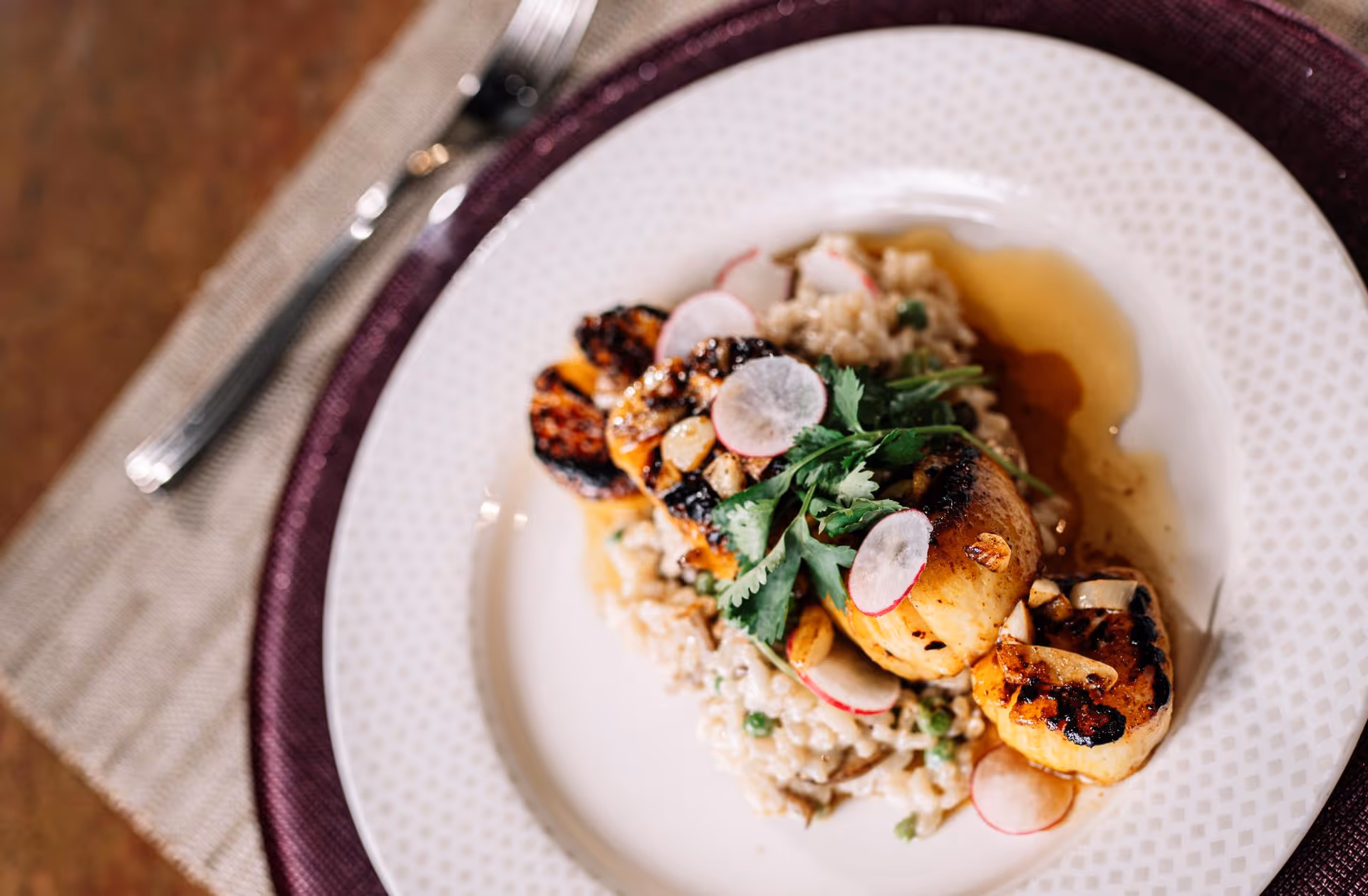 Close-up of grilled scallops served on risotto with radish slices and herbs on a white plate set on a purple charger.
