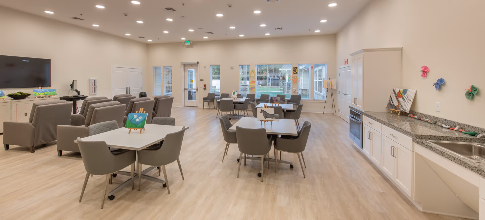 A spacious, well-lit common area in a senior living facility featuring multiple tables with chairs, a seating area with armchairs facing a large wall-mounted TV, and a kitchenette with white cabinets and a granite countertop. The room has light wood flooring and large windows letting in natural light.