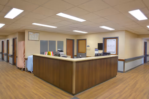 Reception desk area in a nursing and rehabilitation facility with wood paneling, overhead fluorescent lighting, and multiple doors leading to other rooms along the hallway.