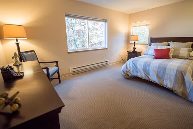 A bright bedroom with a large window letting in natural light. The room features a bed with striped bedding and a red accent pillow, a nightstand with a lamp, a wooden chair, and a dresser with decorative items. The walls are painted light beige and the floor is carpeted.