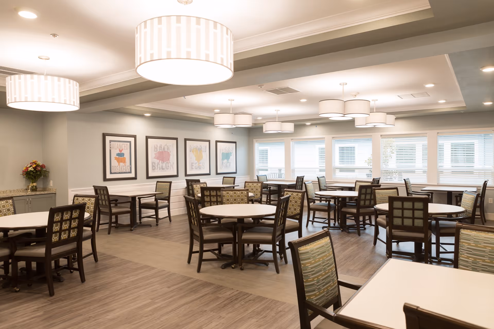 A bright and spacious dining room with multiple round and square tables surrounded by wooden chairs with patterned cushions. The room features large windows with blinds, modern ceiling lights, framed artwork on the walls, and a small counter with a flower arrangement in the corner.