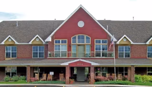 Front exterior view of a two-story assisted living facility building with a red and brick facade, multiple windows, a covered entrance with rocking chairs, and an American flag on a flagpole.