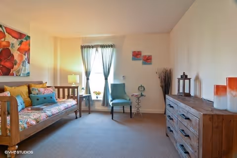 A cozy living room with a wooden daybed adorned with colorful cushions and a floral cover, a teal upholstered chair next to a window with light blue curtains, a wooden dresser with decorative items including candles and a lantern, and wall art featuring red flowers.