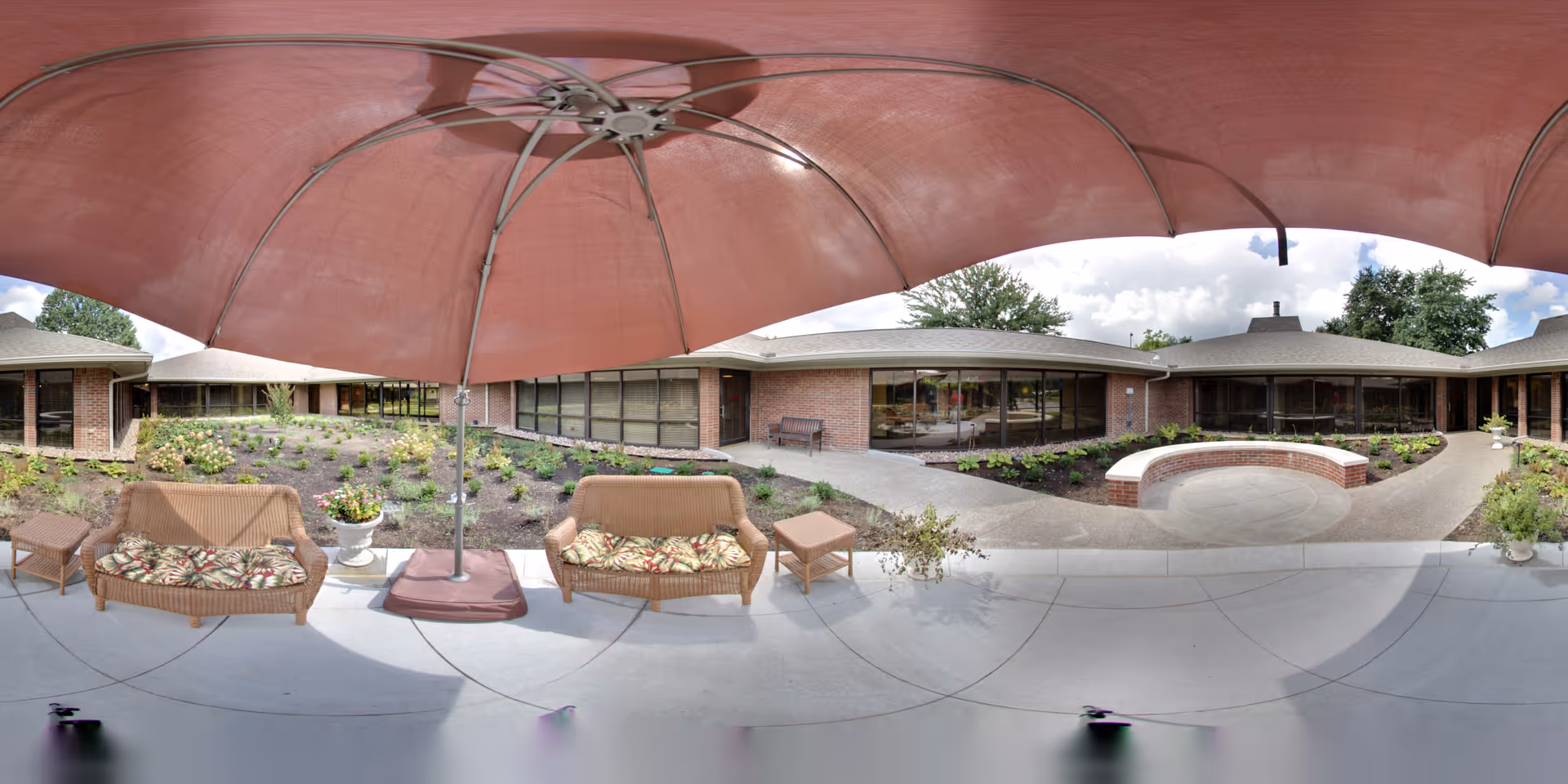 Outdoor courtyard with wicker seating under a large red umbrella, surrounded by a single-story brick building and landscaped plantings.