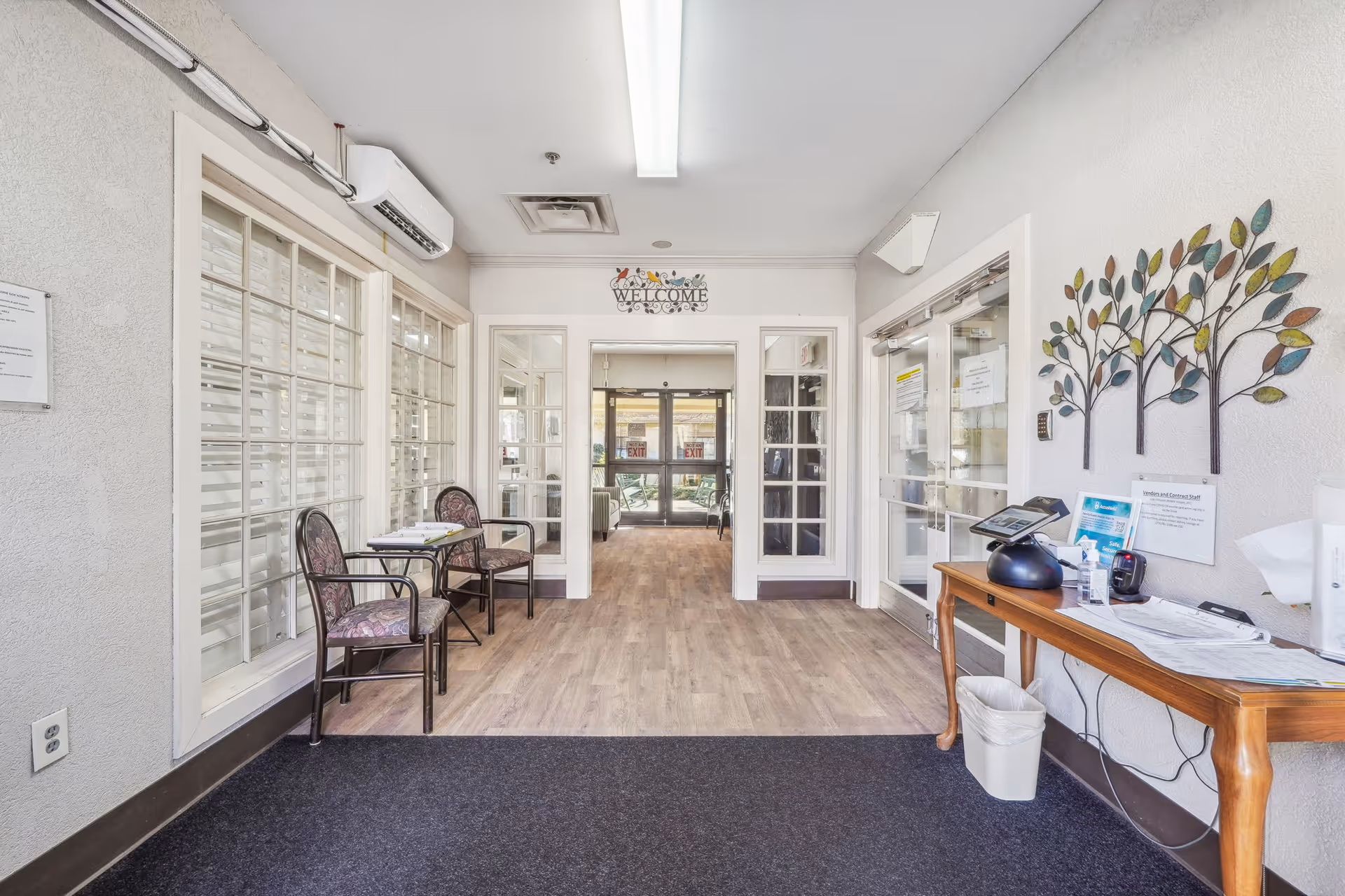 Entrance area of a facility with two chairs and a small table on the left side, a wooden table with papers and a phone on the right side, decorative wall art shaped like trees, and glass doors leading to another room with exit signs visible in the background.