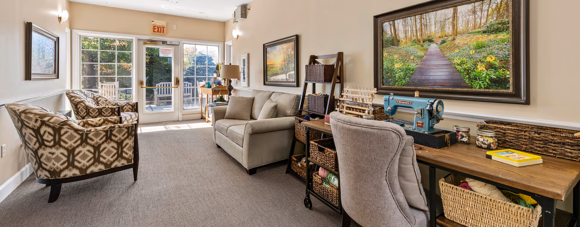 A cozy interior common area with patterned armchairs, a beige sofa, a wooden desk with a vintage sewing machine, baskets, and a large framed picture of a forest path on the wall. Double glass doors lead outside to a patio with wooden chairs.