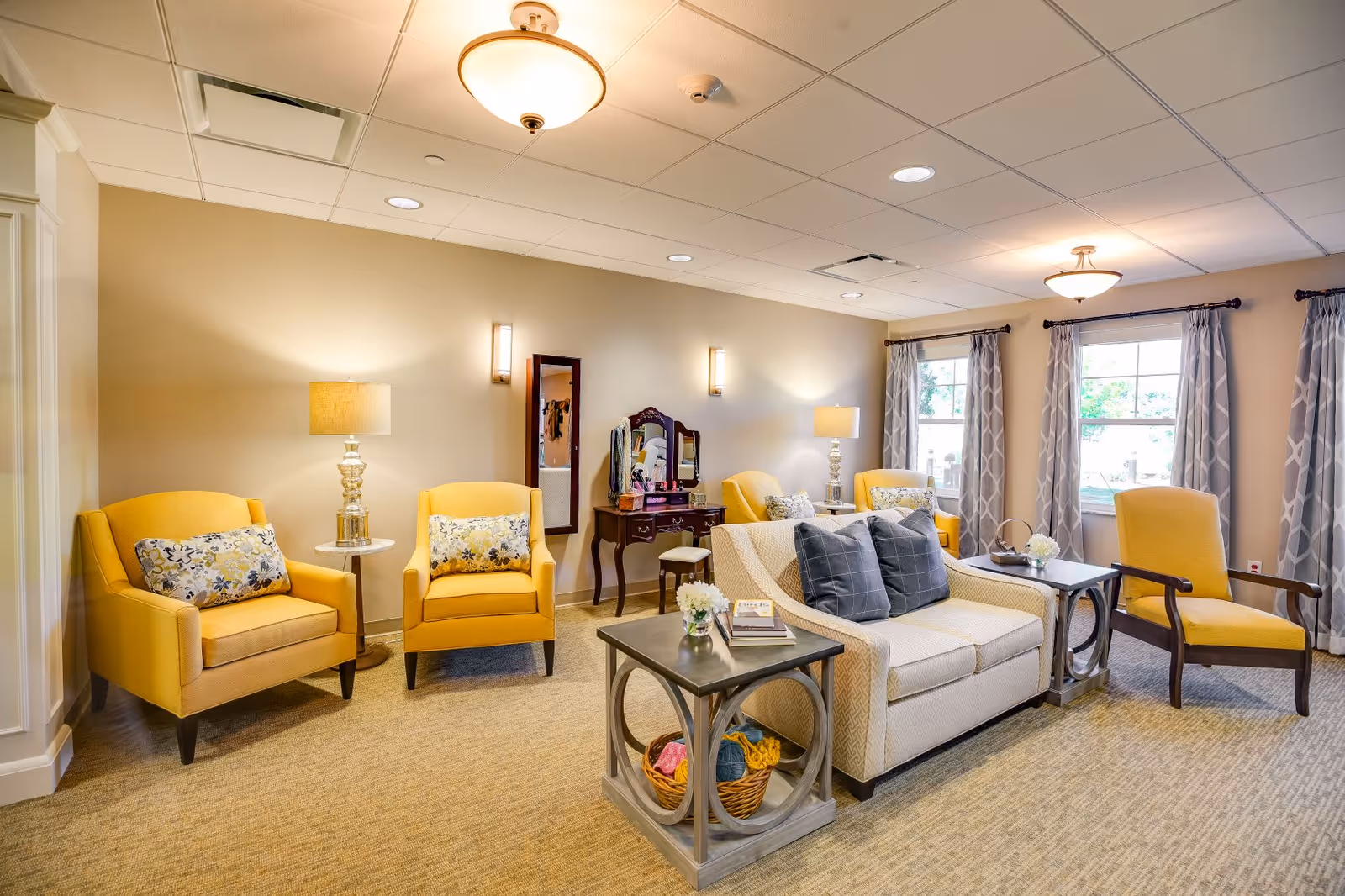 A cozy living room area with beige walls and carpeted floor. The room features two yellow armchairs with floral cushions, a beige loveseat with dark gray pillows, a wooden armchair with yellow cushions, and two side tables with small flower arrangements. There are two large windows with patterned gray curtains allowing natural light to enter. A wooden vanity with a mirror and a small stool is placed against the wall, and the room is softly lit by ceiling lights and table lamps.