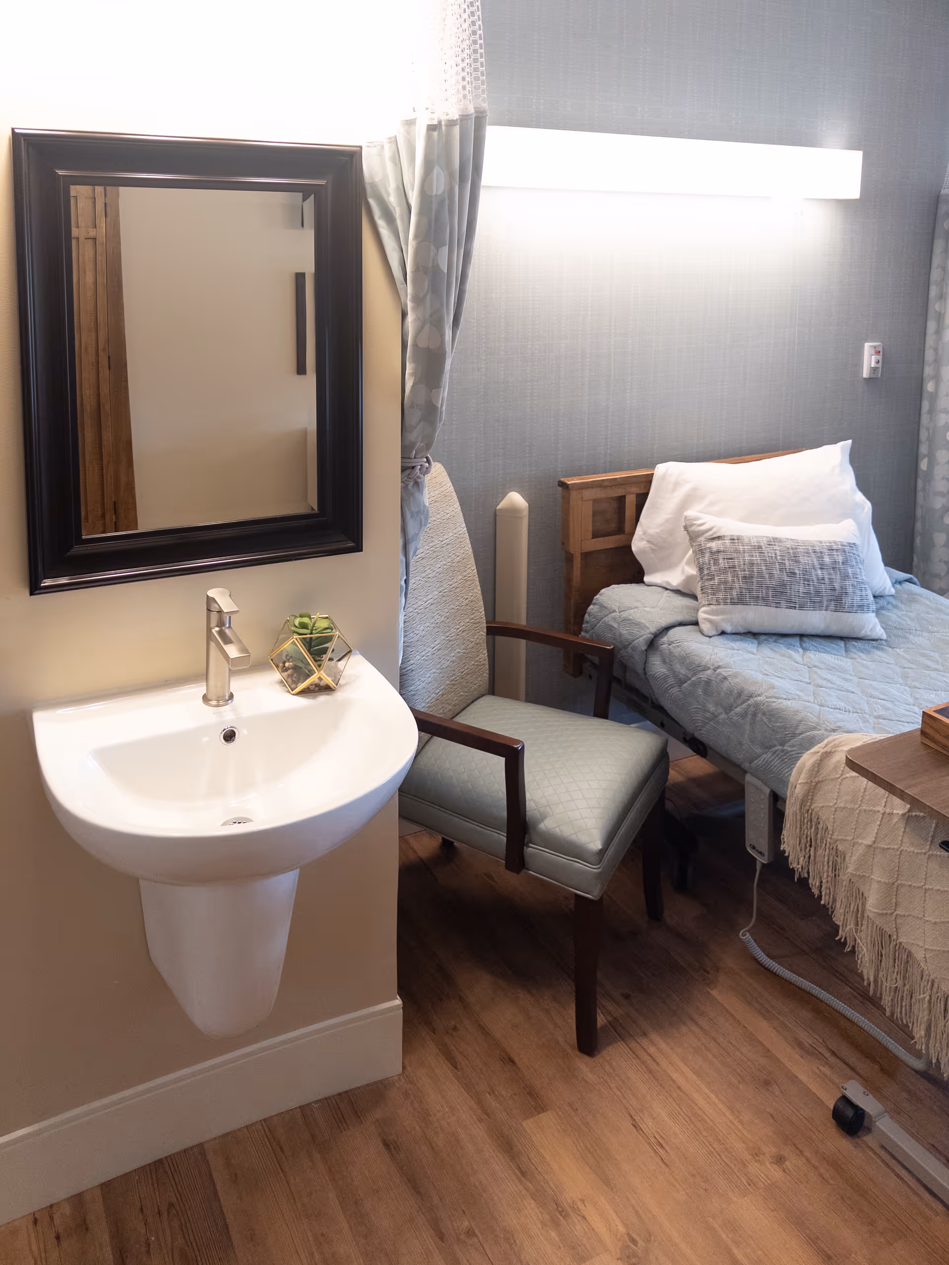 A furnished senior bedroom featuring a bed with pillows, a chair, a wall-mounted sink and mirror, and wood flooring.