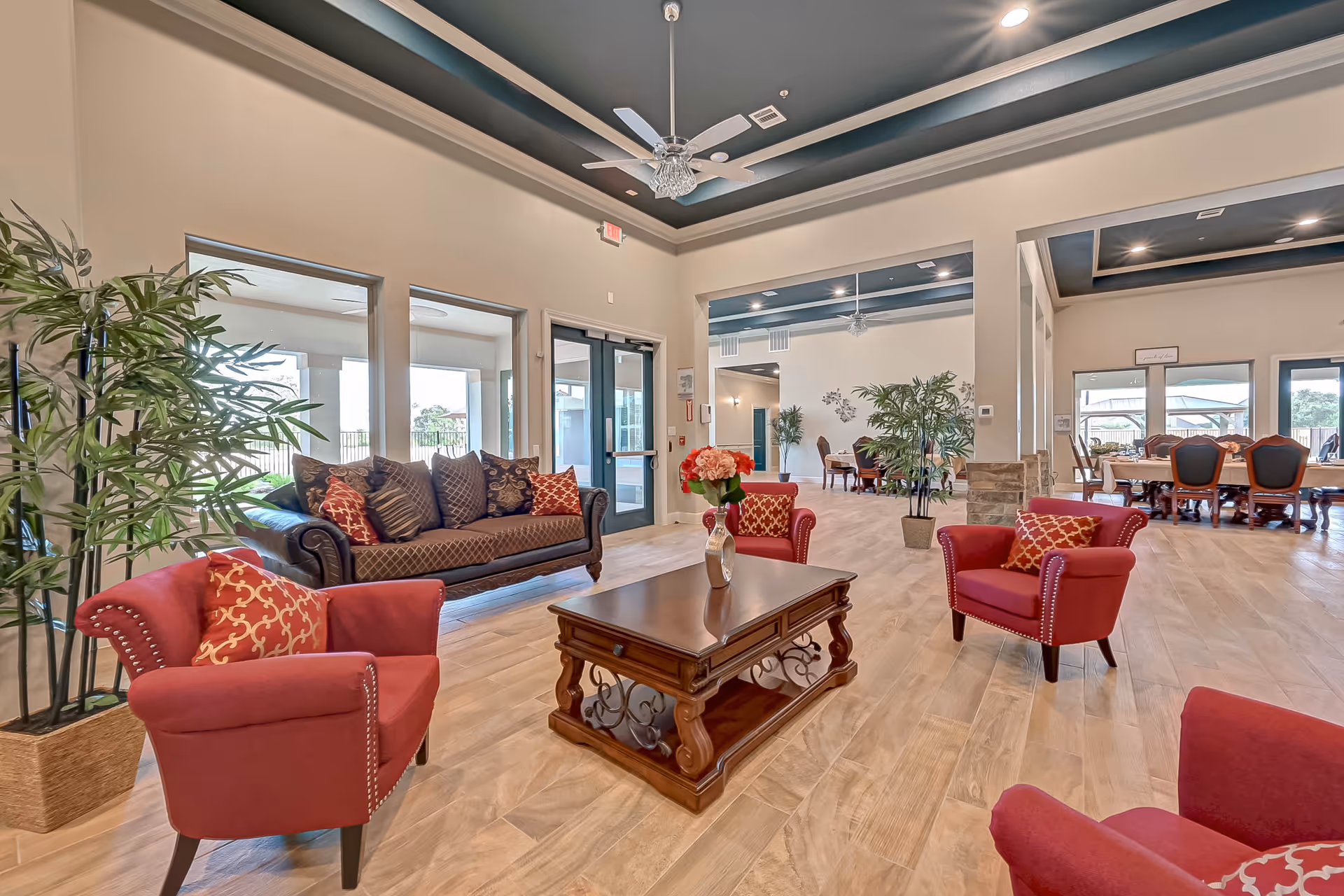 Spacious senior living lounge with red armchairs, a patterned sofa, wooden coffee table, plants, and a dining area visible in the background.