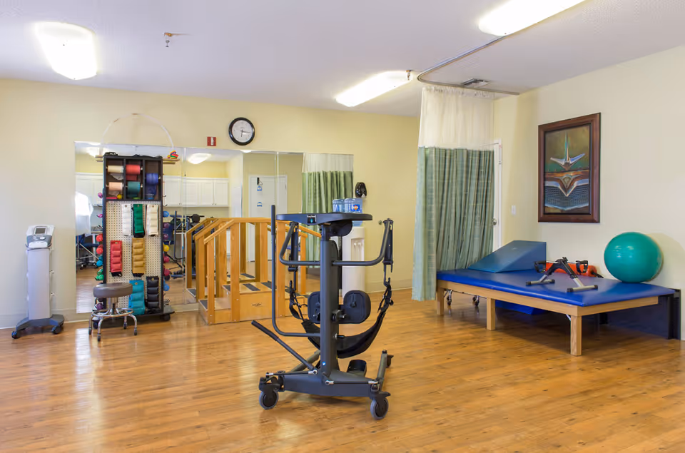 A physical therapy room with wooden flooring, a parallel bar setup for walking exercises, a rack holding various colored exercise bands, a blue therapy table with exercise equipment and a green exercise ball on it, a wall clock, a framed artwork, and a privacy curtain in the corner.
