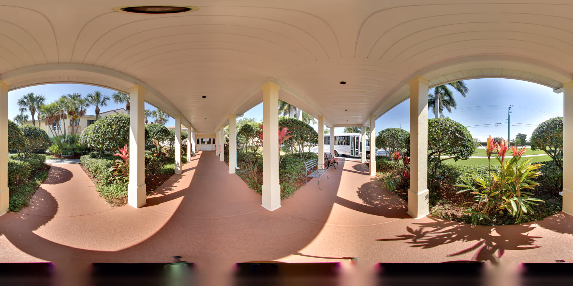 Covered outdoor walkway with white columns and a curved ceiling, surrounded by well-maintained bushes, palm trees, and flowering plants. There are benches along the walkway and a white vehicle parked in the distance under a clear blue sky.