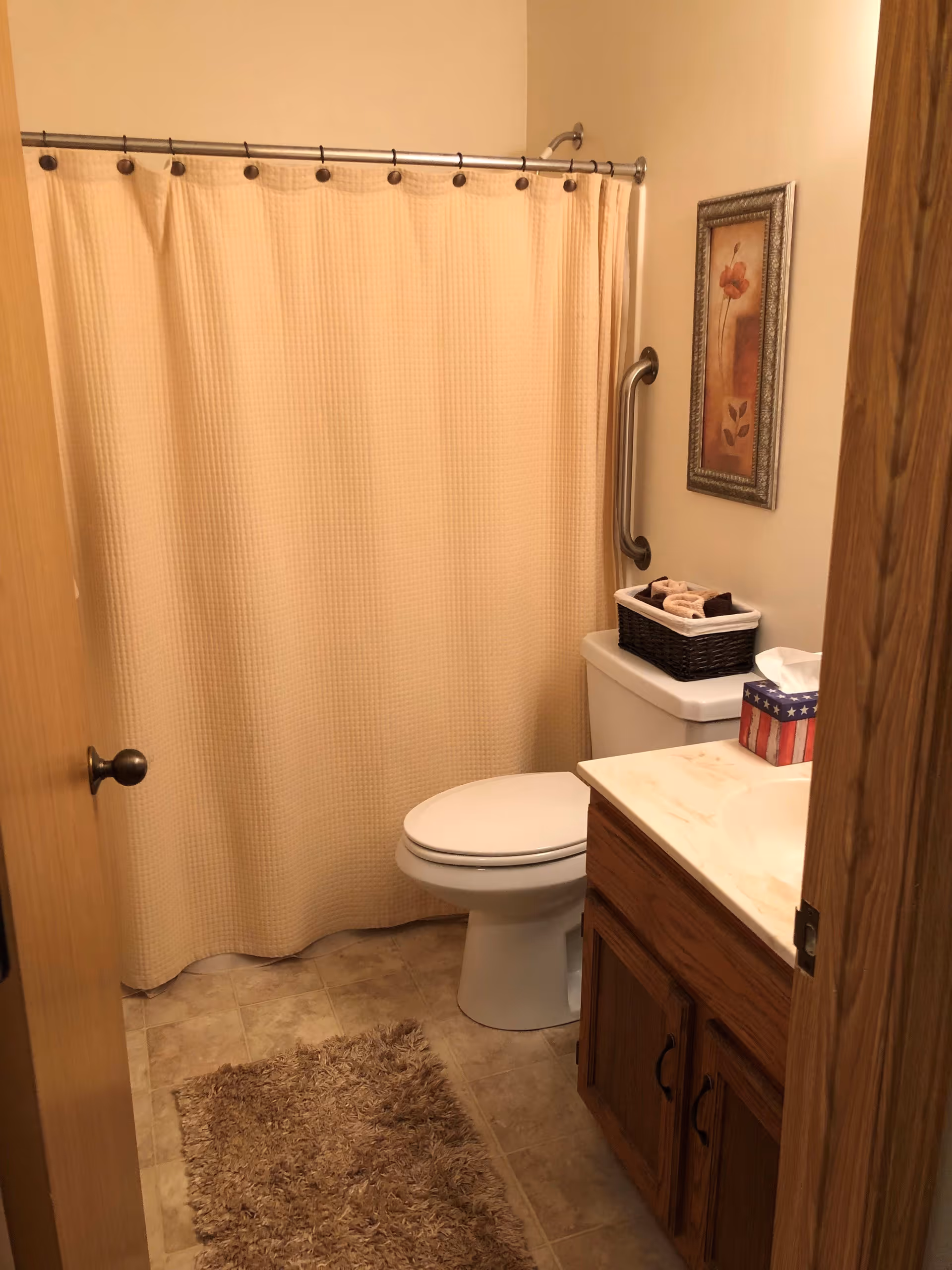 A small bathroom with a beige shower curtain, a white toilet, a wooden vanity with a marble countertop, a basket with towels on the toilet tank, a tissue box with an American flag design, a brown shaggy rug on the tiled floor, and a framed floral picture on the wall.