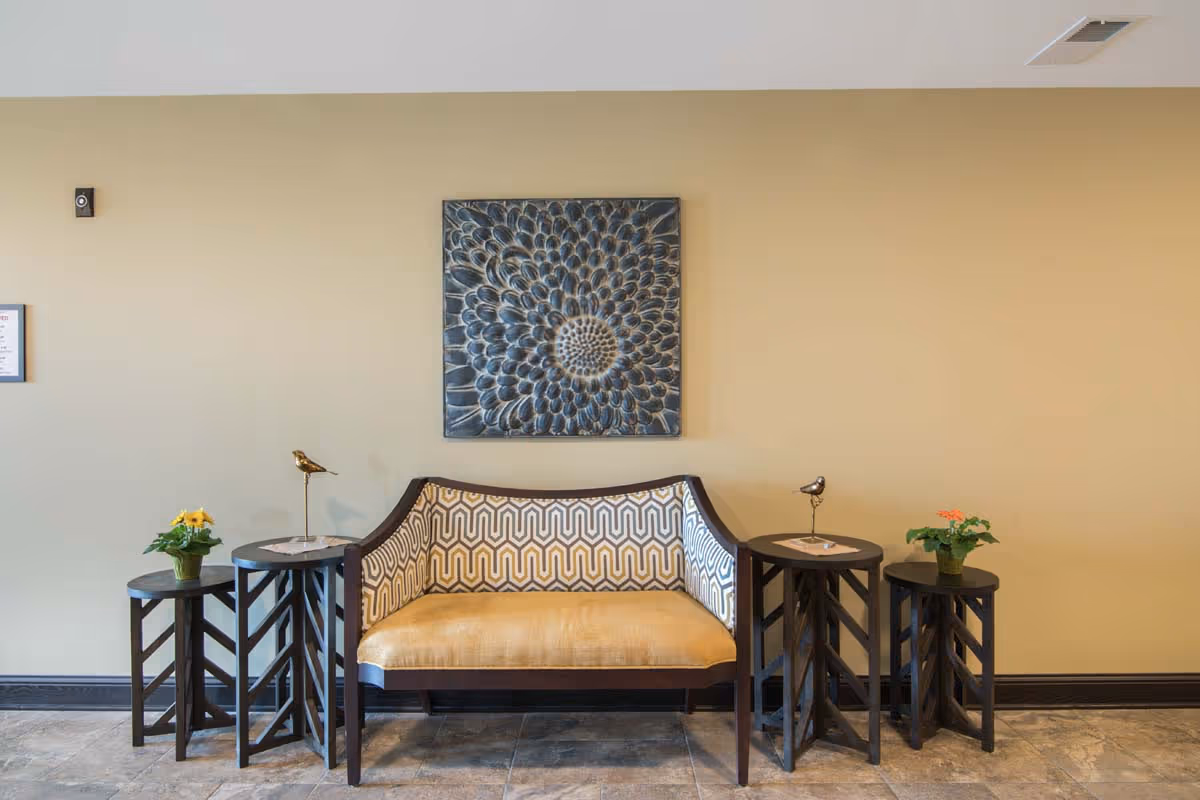 Upholstered bench with a patterned back is flanked by small wooden side tables holding potted plants and decorative bird figurines beneath a large floral wall art in a reception area.