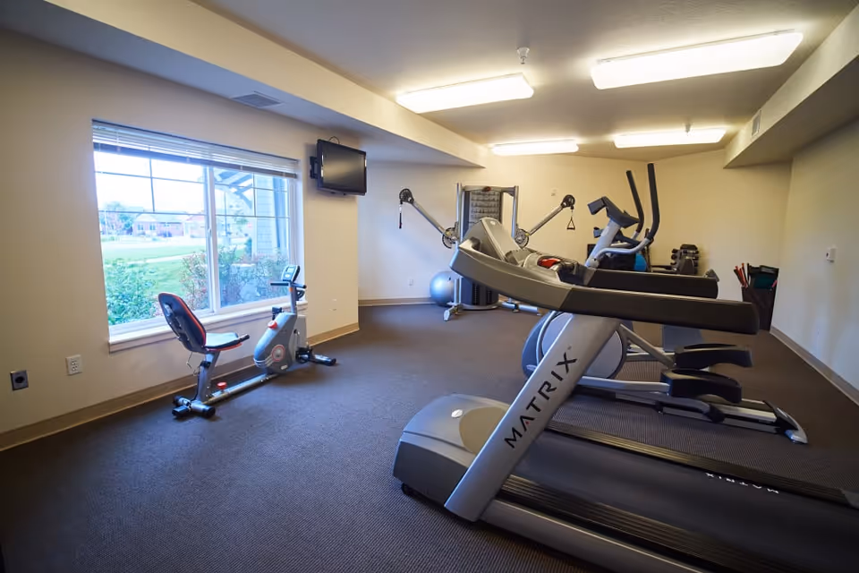 Exercise room with treadmills, a recumbent bike, an elliptical and a strength machine, plus a window and wall-mounted TV.