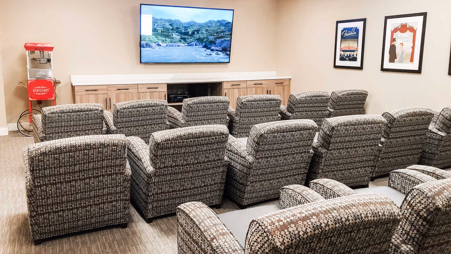 A small theater room with two rows of patterned recliner chairs facing a wall-mounted flat screen TV. There is a red popcorn machine on the left side and three framed vintage-style posters on the right wall. The room has beige walls and carpeted flooring.