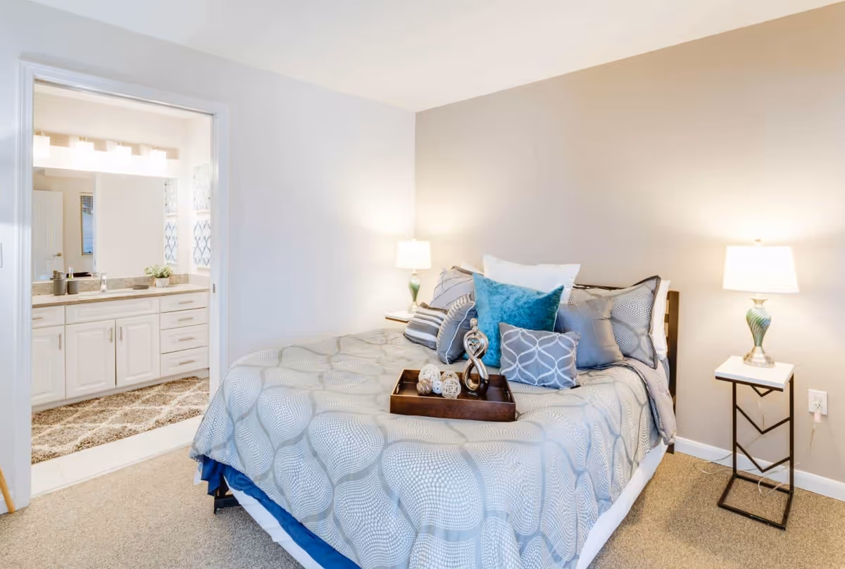 A cozy bedroom with a neatly made bed featuring multiple pillows and a decorative tray on top. Two bedside tables each hold a lamp with a green and white base. The room has beige walls and carpeted flooring. An open doorway reveals a bathroom with a large mirror, white cabinets, and a granite countertop.