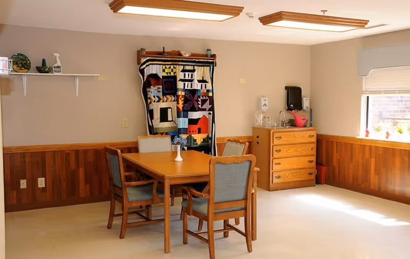 Small communal dining/activity room with a wooden table and four chairs, a colorful quilt hanging on the wall, and a dresser sink by a window.