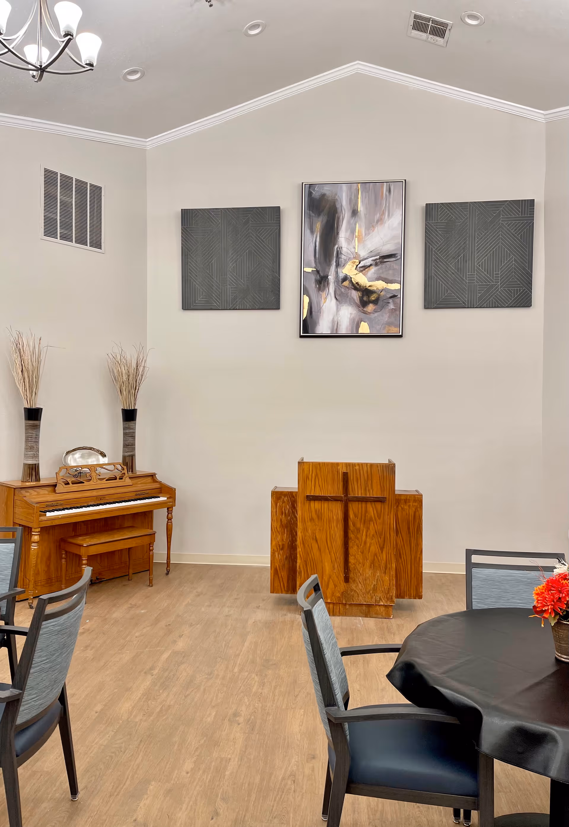 Interior room with light-colored walls and wooden flooring featuring a wooden piano with two tall vases holding dried plants on top, a wooden podium with a cross on the front, three abstract paintings on the wall above the podium, and a round table with a black tablecloth and chairs around it.