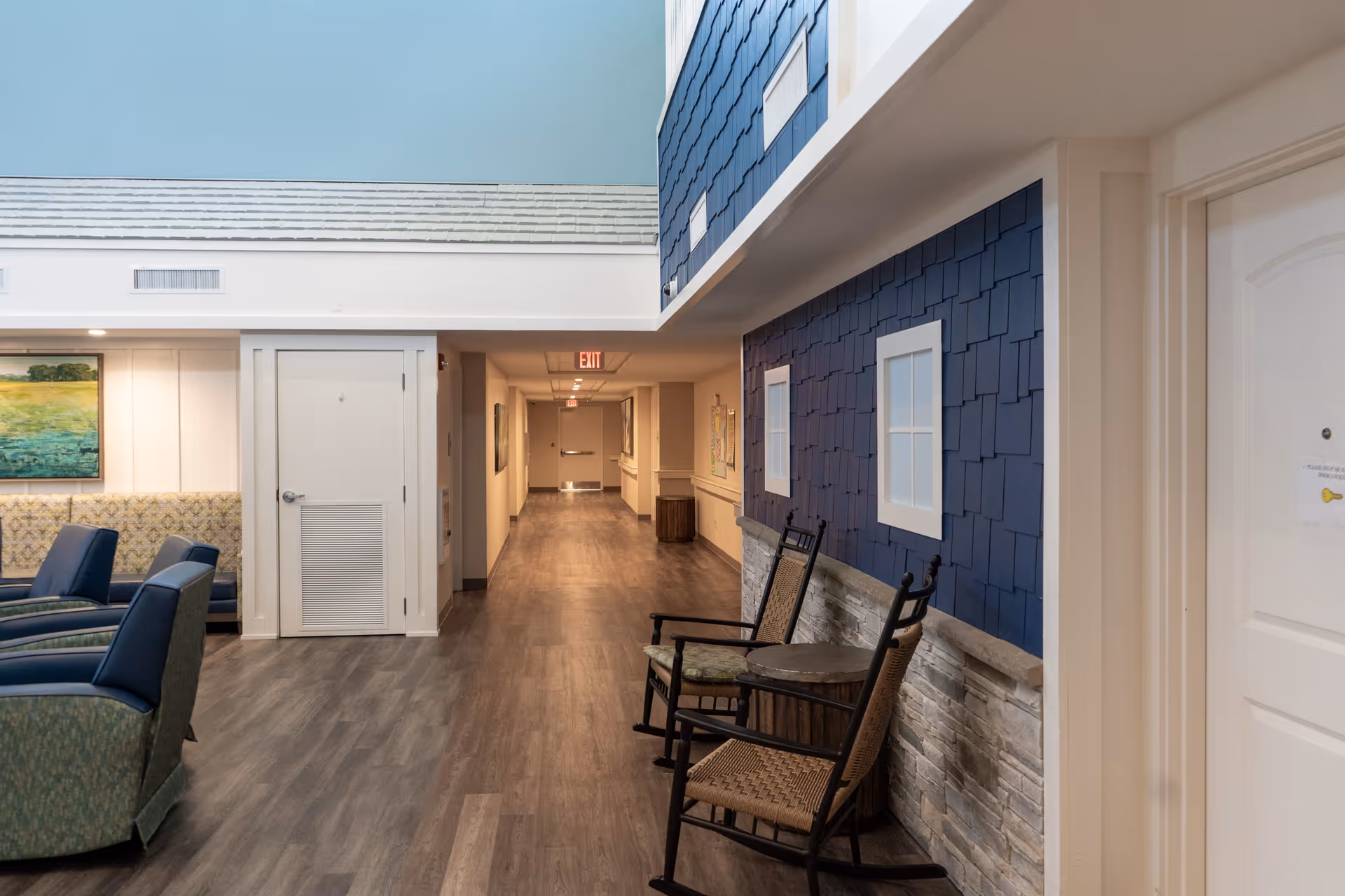 Interior hallway of The Landing of Panama City Beach facility with wood flooring, blue shingle-style wall with two white-framed windows, two wooden rocking chairs and a small round table. On the left side, there are upholstered chairs and a bench with a landscape painting above it. The hallway extends to a door with an exit sign above it.