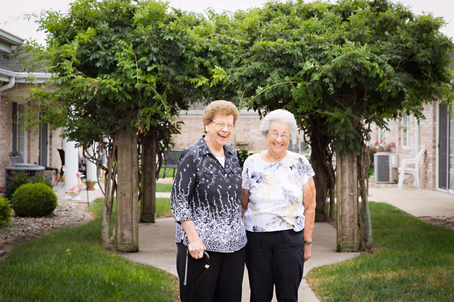 Two elderly women standing and smiling together under a green leafy arbor in an outdoor garden area of a senior living facility.