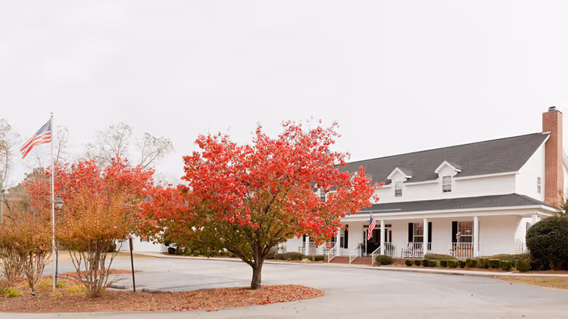 A large white two-story building with a dark roof and a front porch with rocking chairs. In front of the building, there is a tree with bright red autumn leaves and some smaller trees with orange leaves. An American flag is flying on a flagpole near the trees. The scene appears to be in a parking or driveway area with some bushes along the building.