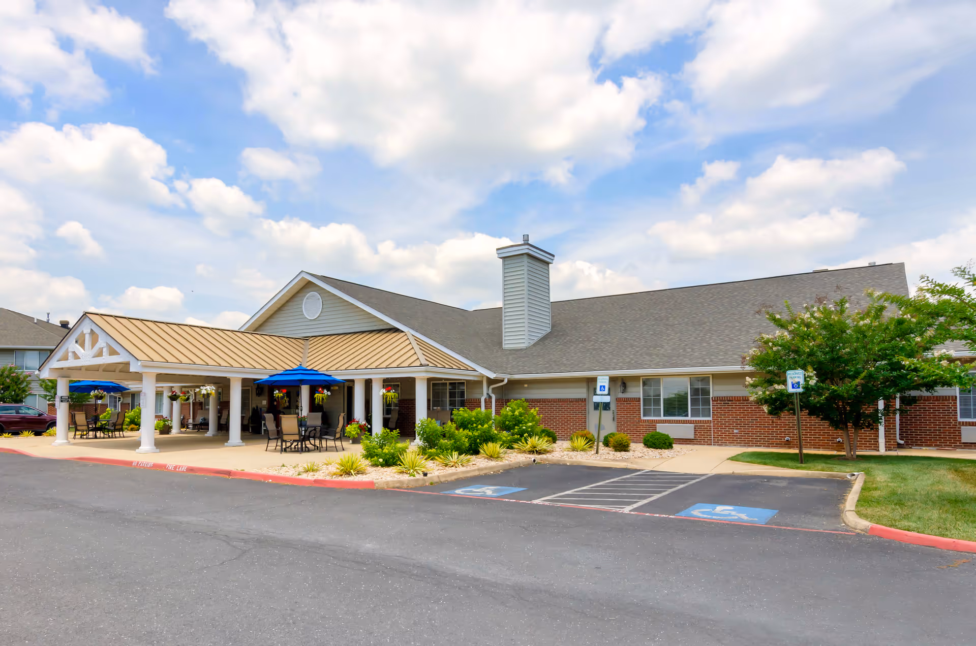 Exterior view of a single-story brick and siding building with a covered entrance featuring white columns and a tan roof. There are outdoor tables with blue umbrellas and hanging flower baskets. The parking lot in front has designated handicapped parking spaces. The sky is partly cloudy.