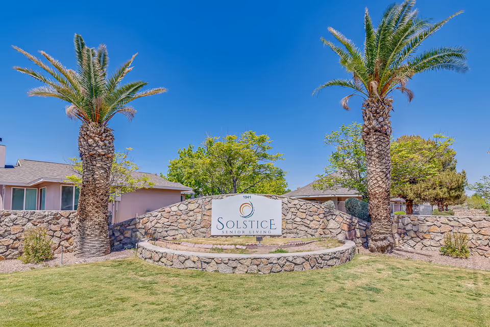 Outdoor view of the entrance sign for Solstice Senior Living at Rio Norte, featuring a stone wall with a white sign, two tall palm trees, green grass, and clear blue sky.