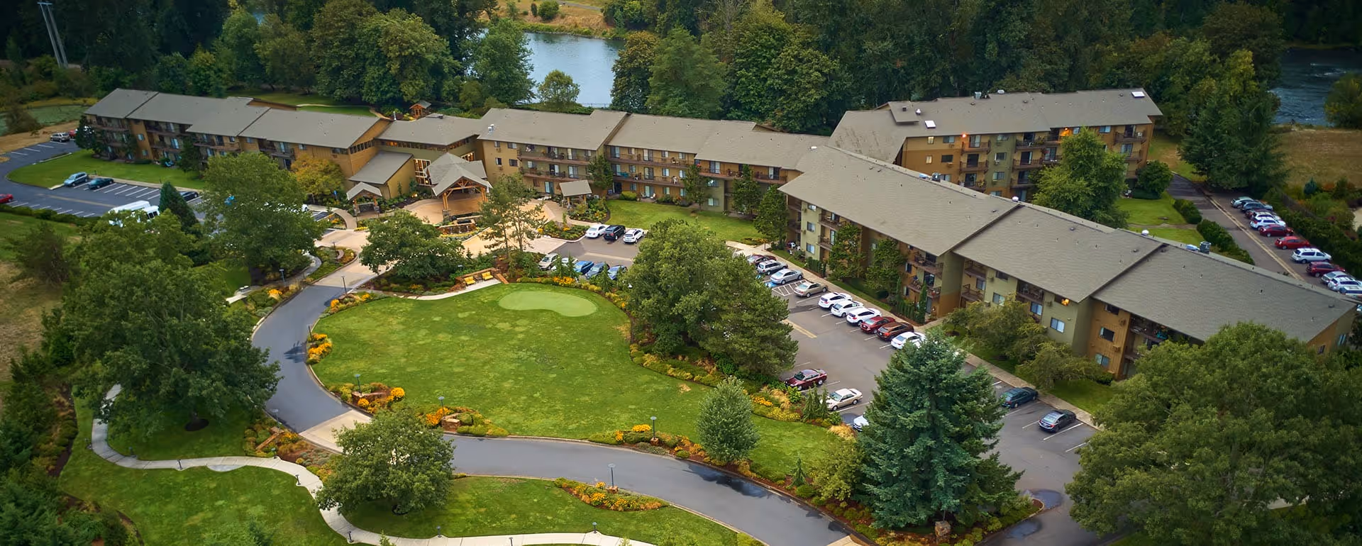 Aerial view of Willamette Oaks senior living facility showing multiple connected buildings surrounded by trees, parking lots, and landscaped green areas with a small pond in the background.