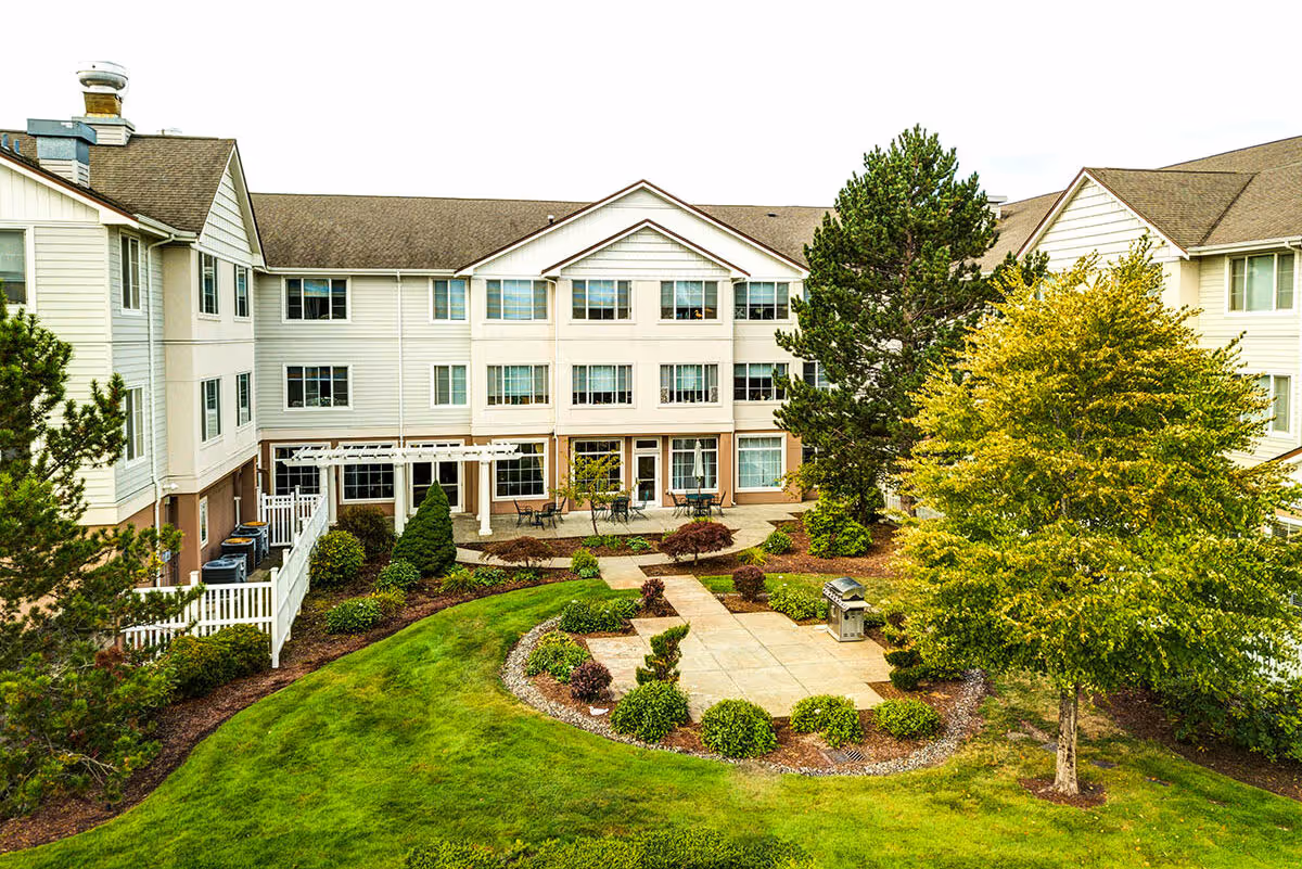 Three-story senior living building overlooking a landscaped central courtyard with patio seating and trees.
