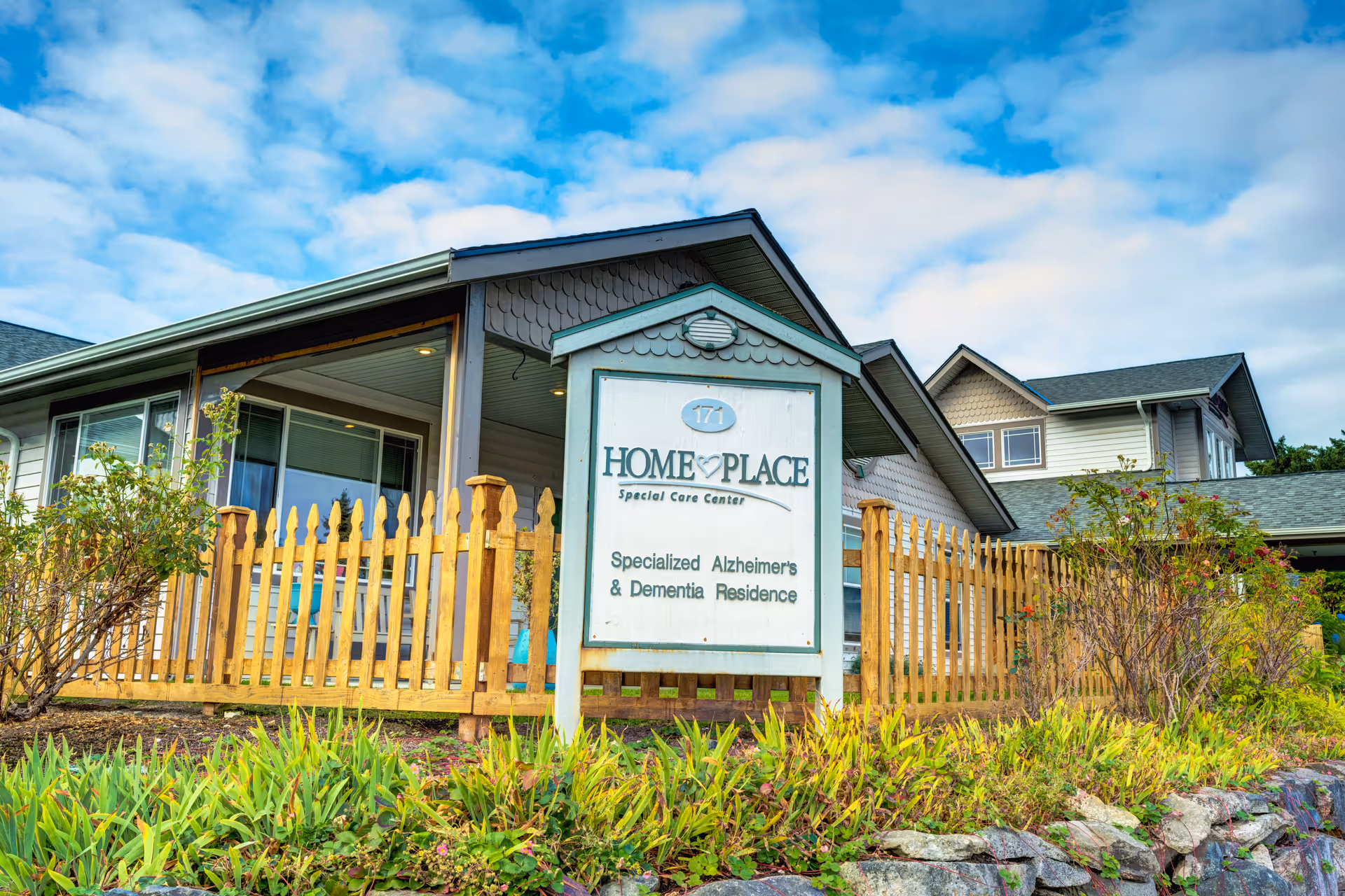 Exterior view of HomePlace Special Care Center at Oak Harbor, showing a wooden picket fence, a sign indicating it is a specialized Alzheimer's and dementia residence, and part of the building under a partly cloudy sky.