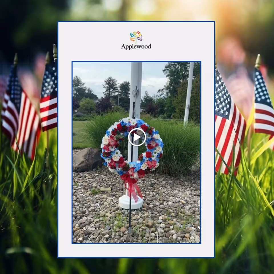 A patriotic floral wreath with red, white, and blue flowers displayed on a stand in a landscaped area with rocks and greenery. The background shows trees and a cloudy sky. The image is framed with the Applewood logo at the top and American flags blurred in the foreground.