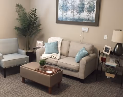 A cozy living room area with a beige loveseat adorned with two blue pillows and a white knitted throw blanket. Next to it is a light blue armchair. In front of the loveseat is a rectangular ottoman with a wooden tray holding a small plant and some glasses. A tall green plant is placed in the corner, and a framed picture of trees hangs on the beige wall above the loveseat. To the right, there is a small side table with a lamp, framed photo, and some books.