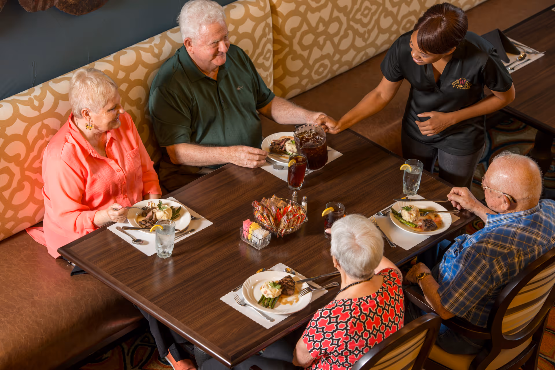 Four elderly people seated around a dining table enjoying a meal with plates of food and glasses of water with lemon. A staff member in a black uniform with a Watercrest logo is serving a pitcher of iced tea to one of the men. The seating includes a patterned cushioned bench and chairs.