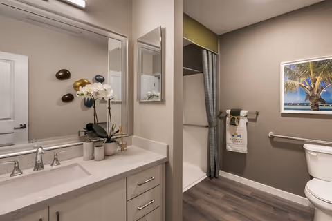 A modern bathroom featuring a large mirror above a white countertop with a sink, decorated with a white orchid plant and small containers. The bathroom has a toilet with a grab bar beside it, a towel rack with folded towels, and a shower area with a curtain. The walls are painted beige, and there is a framed picture of a palm tree on the wall.