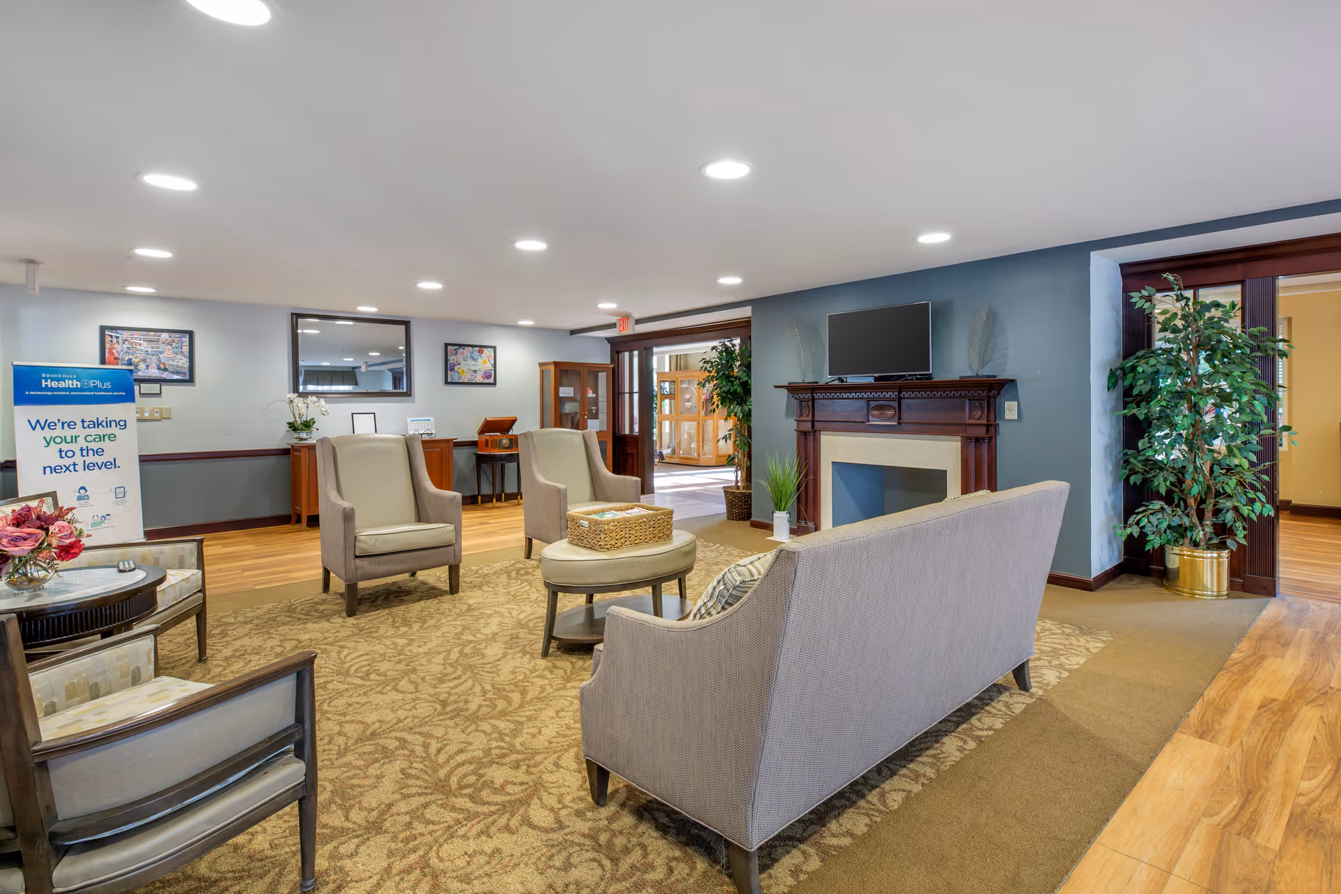 A well-lit senior living facility common area with beige armchairs and a sofa arranged around a small round table on a patterned carpet. There is a wooden fireplace with a flat-screen TV mounted above it, potted plants, framed artwork on the walls, and a sign promoting Brookdale HealthPlus in the background.
