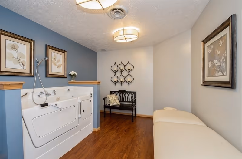 A room with a walk-in bathtub on the left side, decorated with two framed botanical prints above it. There is a black wooden bench with a decorative pillow and a wall-mounted candle holder with multiple candles on the back wall. On the right side, there is a cushioned massage or treatment table and a framed floral artwork on the wall. The room has wooden flooring and soft lighting from ceiling fixtures.