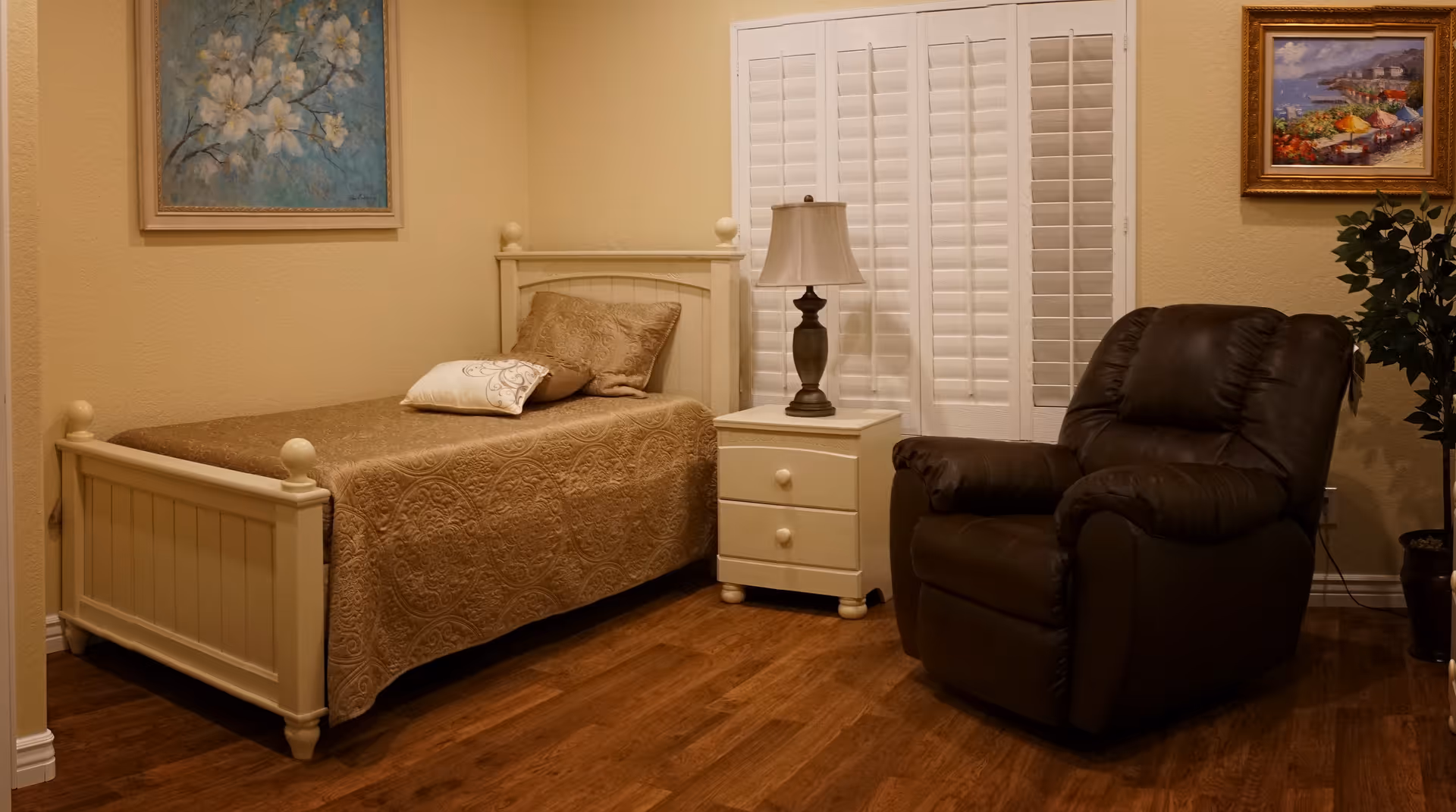 A cozy bedroom with a single bed covered in a brown quilt and two pillows. Next to the bed is a white nightstand with a lamp on it. There is a dark brown recliner chair to the right of the nightstand. The room has wooden flooring, beige walls, white plantation shutters on the window, and two framed paintings on the walls. A potted plant is visible in the corner.
