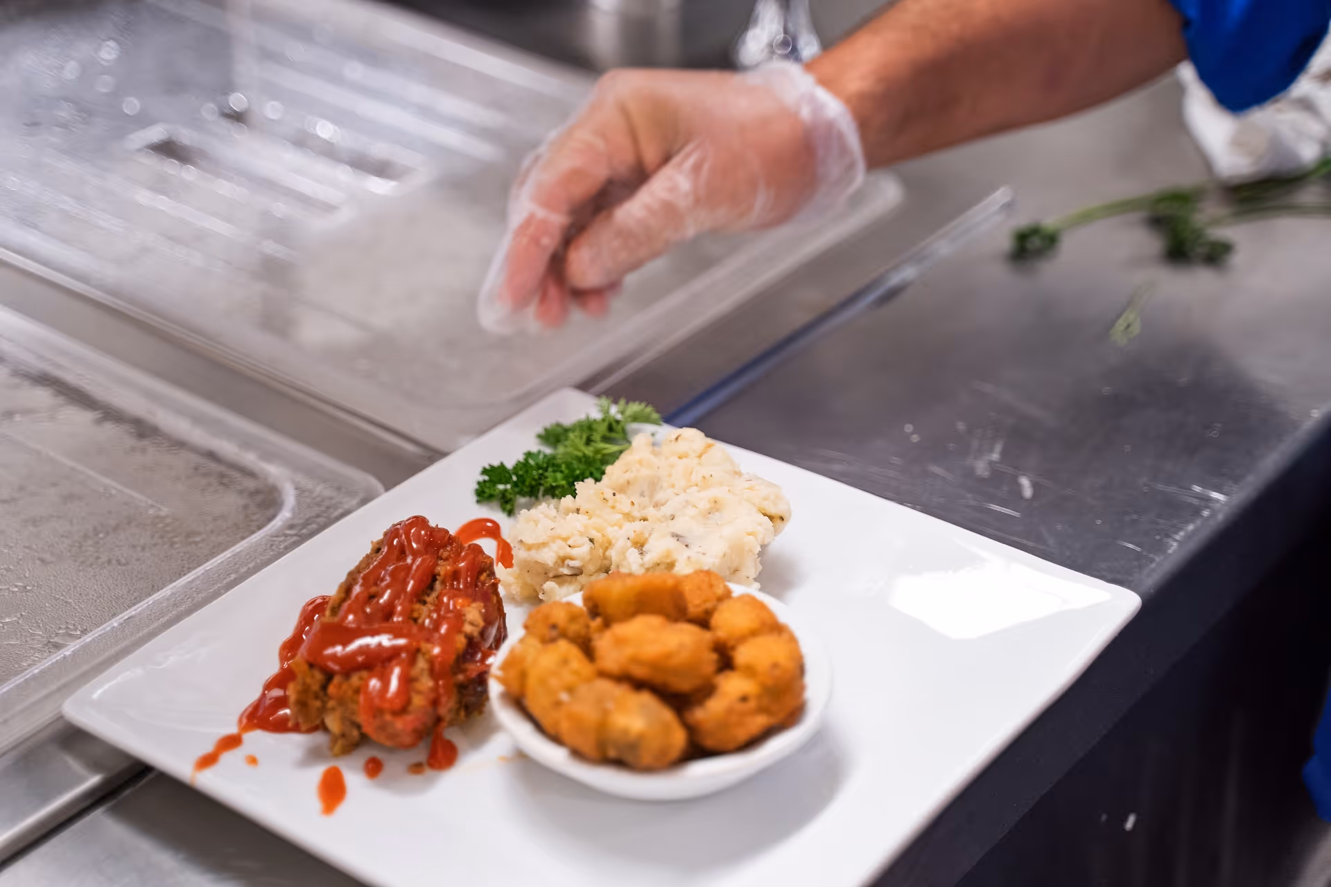 A plate of food on a stainless steel counter with mashed potatoes, fried chicken topped with ketchup, and a small bowl of fried hush puppies. A person wearing a blue sleeve and a disposable glove is placing the plate down.