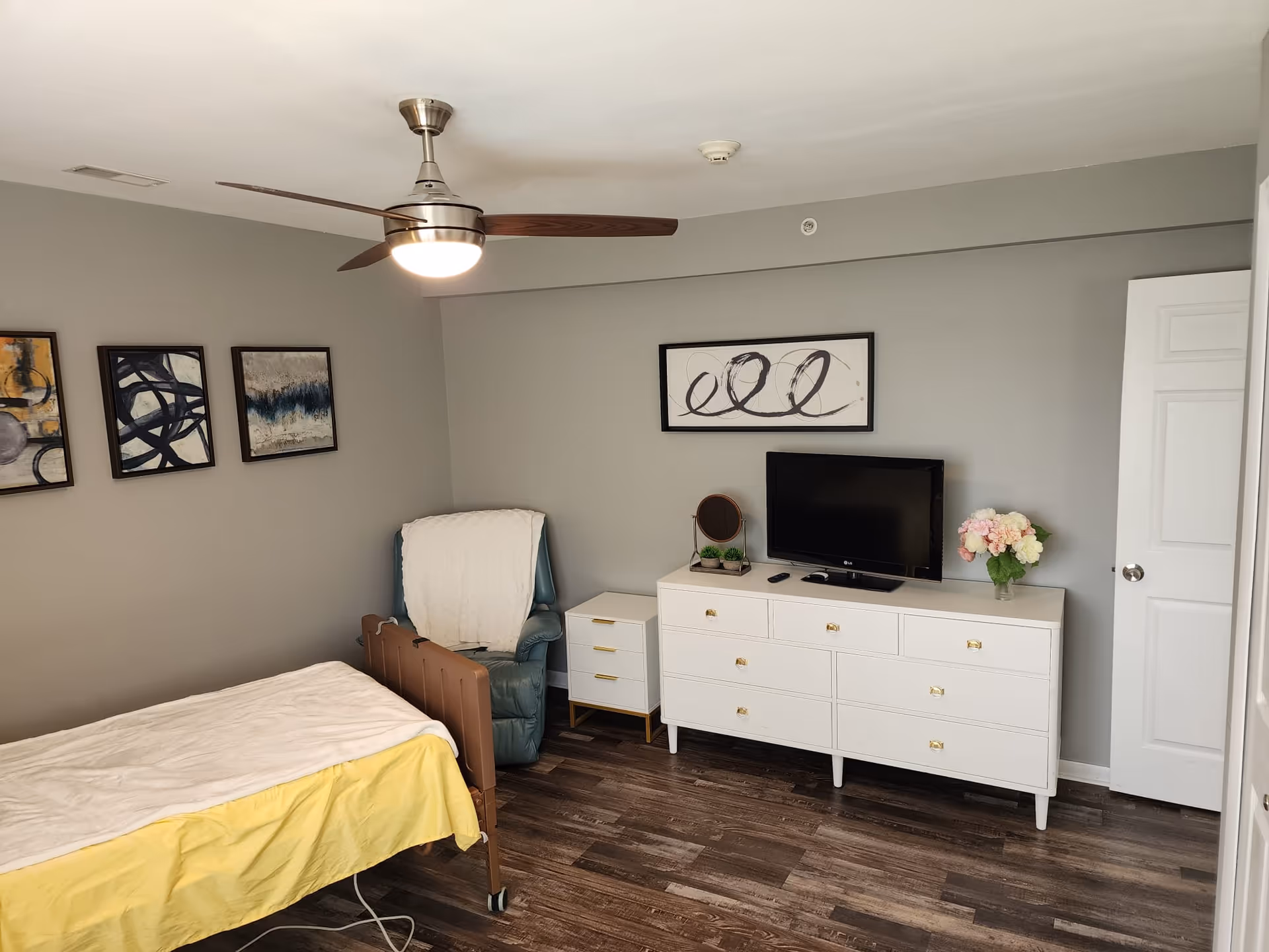 A bedroom with a single bed covered with a yellow and white blanket, a blue armchair with a white throw draped over it, a white dresser with a flat-screen TV, a small mirror, and a vase of flowers. The walls are gray with abstract artwork hanging, and there is a ceiling fan with a light fixture.