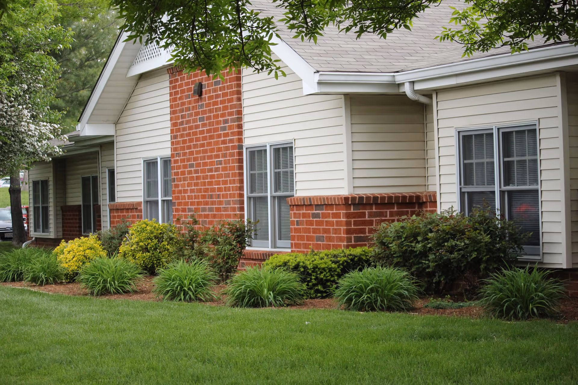Exterior view of a single-story building with beige siding and red brick accents, surrounded by green shrubs, plants, and a well-maintained lawn.