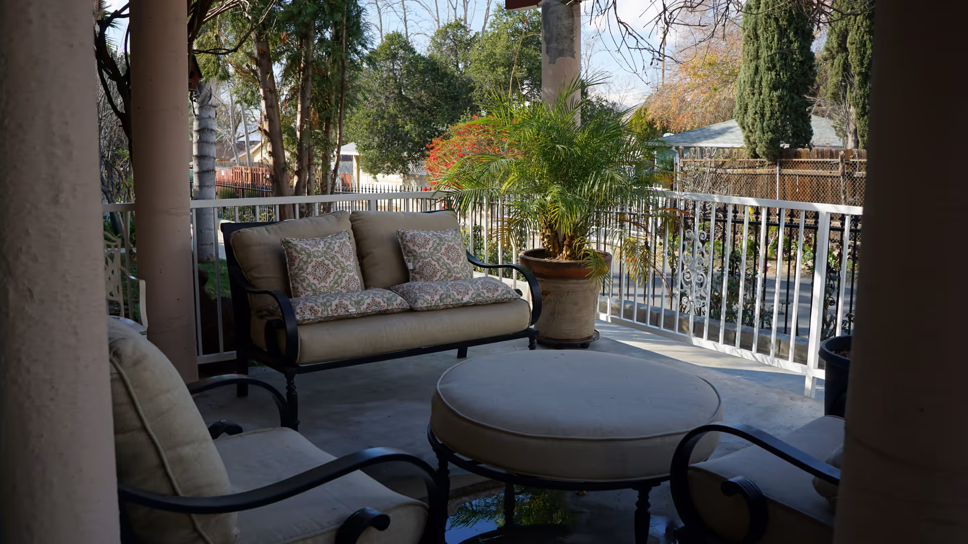 A covered outdoor patio area with cushioned seating including a loveseat, two armchairs, and a round ottoman. There is a large potted plant near the white railing, and trees and houses are visible in the background.
