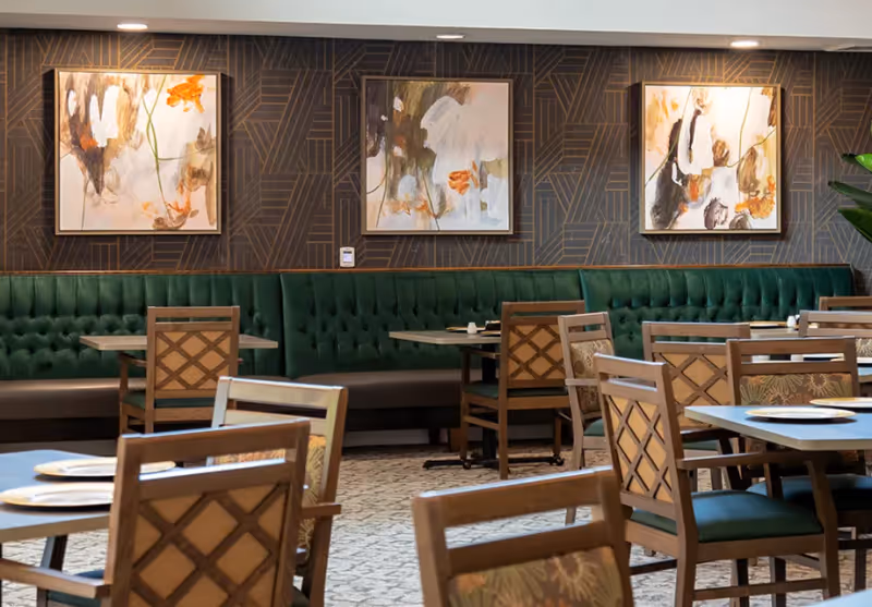 Interior view of a dining room with wooden tables and chairs, green cushioned bench seating along the wall, and three abstract paintings hanging on a dark patterned wall.