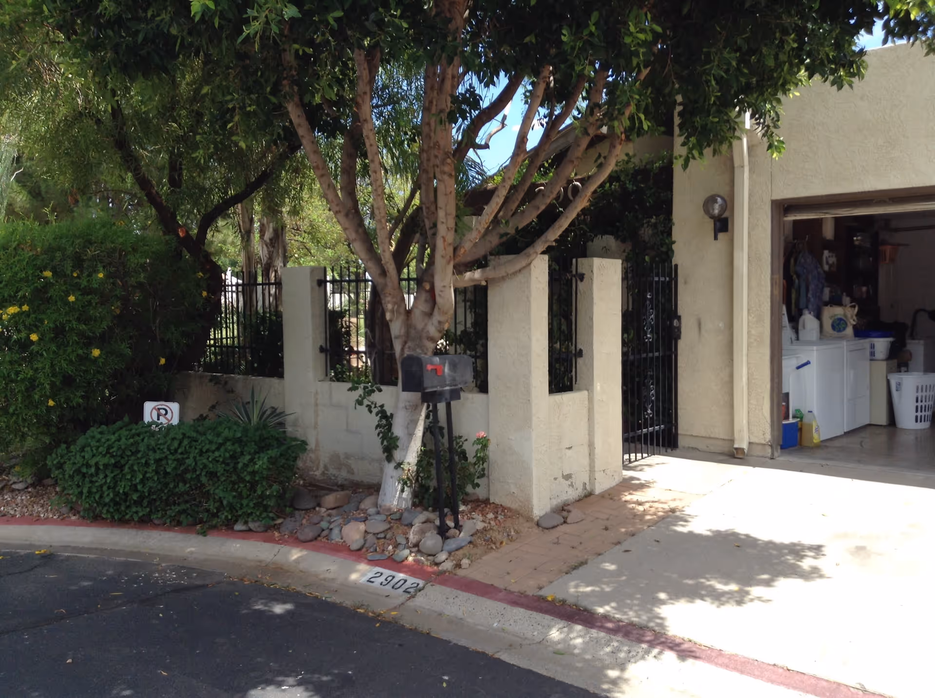 A residential driveway and entrance area with a mailbox mounted on a post near a tree. There is a beige stucco wall with a black metal gate, and an open garage showing laundry appliances and household items inside. The area is shaded by trees and surrounded by greenery, with a curb marked with the number 2902.