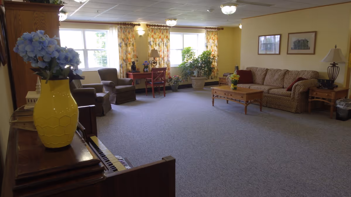 A cozy living room with a patterned sofa, two armchairs, a wooden coffee table with a yellow vase of flowers, a wooden side table with a lamp, and a piano with a yellow vase of blue flowers on top. The room has yellow walls, floral curtains on two windows, and several potted plants near the windows.
