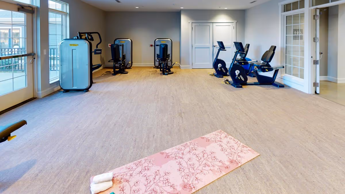 A fitness room with exercise equipment including stationary bikes and weight machines. A pink yoga mat with a rolled white towel is placed on the floor. Large windows and glass doors allow natural light to enter the room.