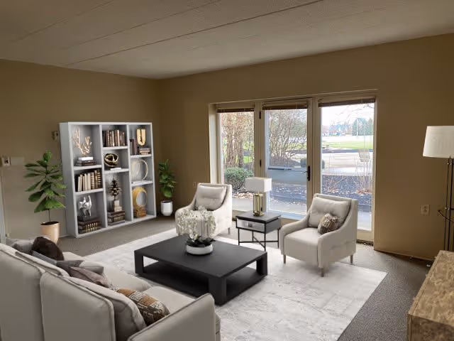 A cozy living room with beige walls and carpeted floor. The room features a light gray sofa with patterned pillows, two matching armchairs, a black coffee table with a white floral centerpiece, and a side table with a lamp. There is a tall bookshelf filled with decorative items and books, two potted plants, and large glass doors leading outside, allowing natural light to fill the space.
