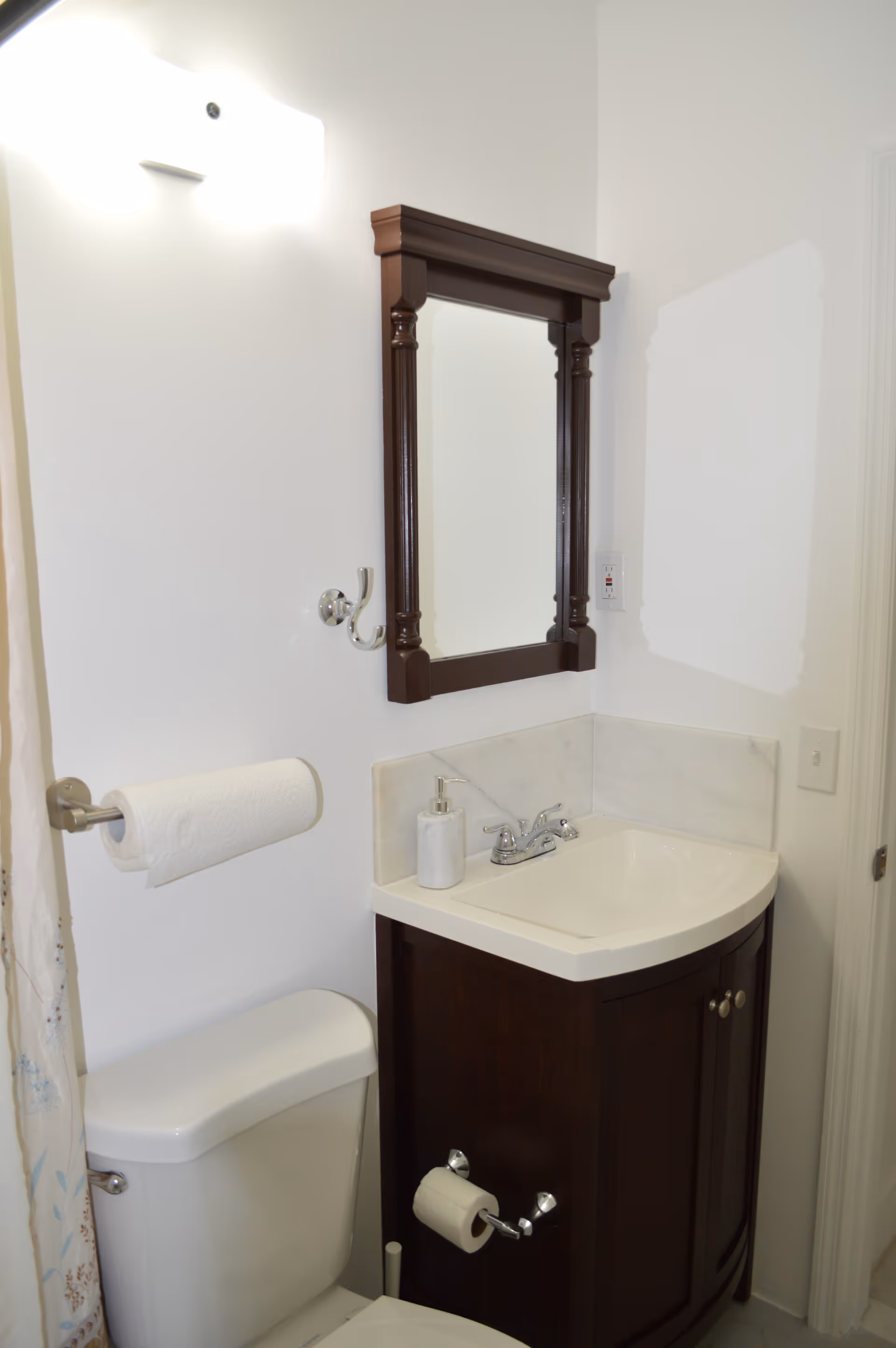 A small bathroom corner featuring a white toilet with a paper towel holder mounted on the wall above it, a dark wood vanity with a white sink and silver faucet, a soap dispenser on the sink, and a dark wood framed mirror mounted on the white wall above the sink. A light fixture is visible above the mirror.