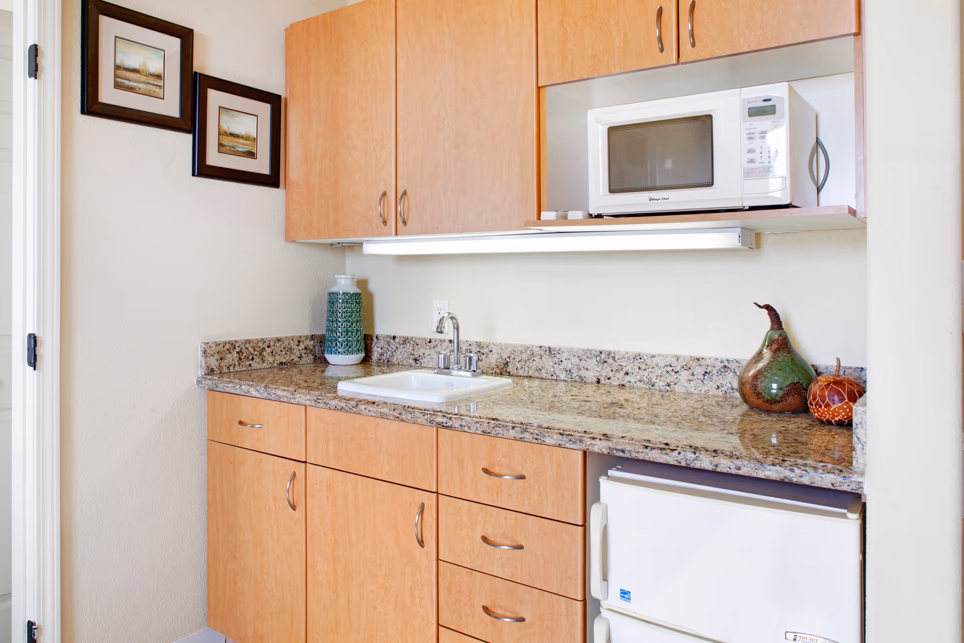 A small kitchen area with light wood cabinets, a granite countertop, a white sink with a faucet, a white microwave on a shelf above the counter, and a small white refrigerator below the counter. There are two framed pictures on the wall to the left and decorative ceramic items on the countertop.