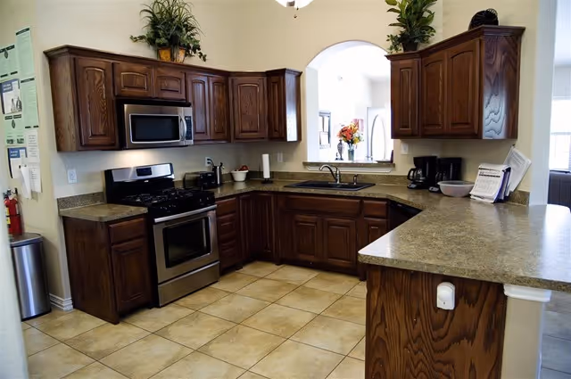 A kitchen with dark wooden cabinets, a stainless steel gas stove and oven, a microwave, a toaster, a coffee maker, and a sink under a pass-through window. The floor is tiled and there are some plants and decorative items on top of the cabinets.