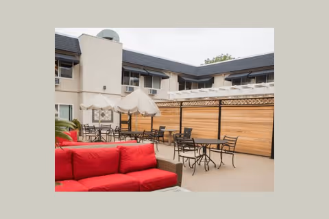Outdoor courtyard with a red patio sofa, tables, chairs and umbrellas in front of a two-story senior living building.